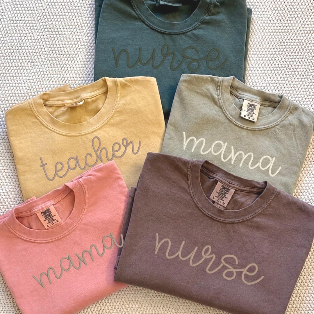Comfort Colors fall t-shirts with embroidered script font reading Mama, Teacher, and Nurse resting on an autumn leaf background.