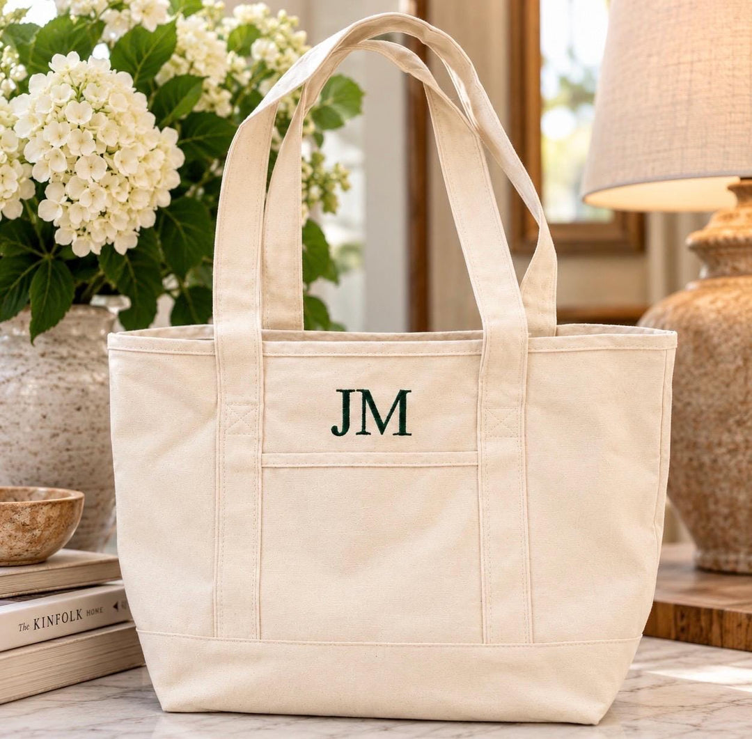 Natural canvas tote bag with dark green embroidered three-letter monogram and front exterior pocket.