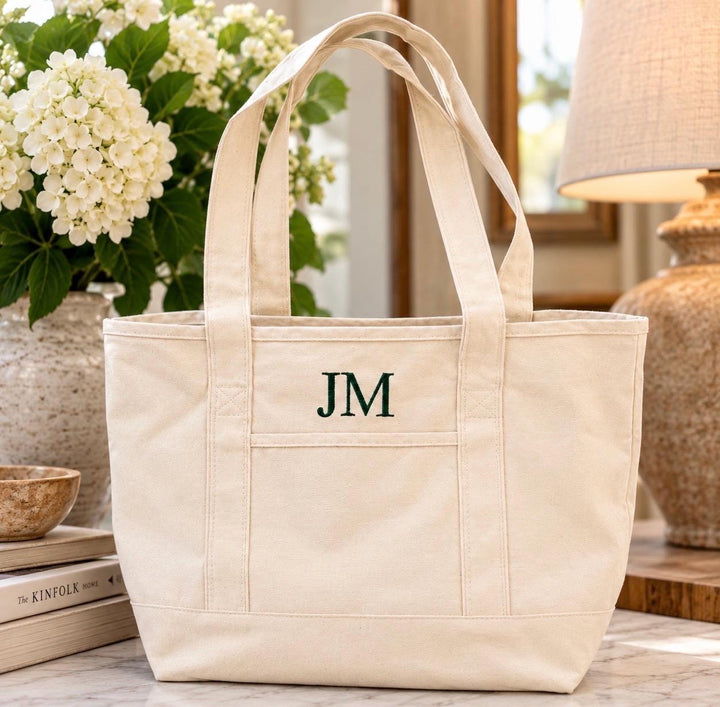 Natural canvas tote bag with dark green embroidered three-letter monogram and front exterior pocket.